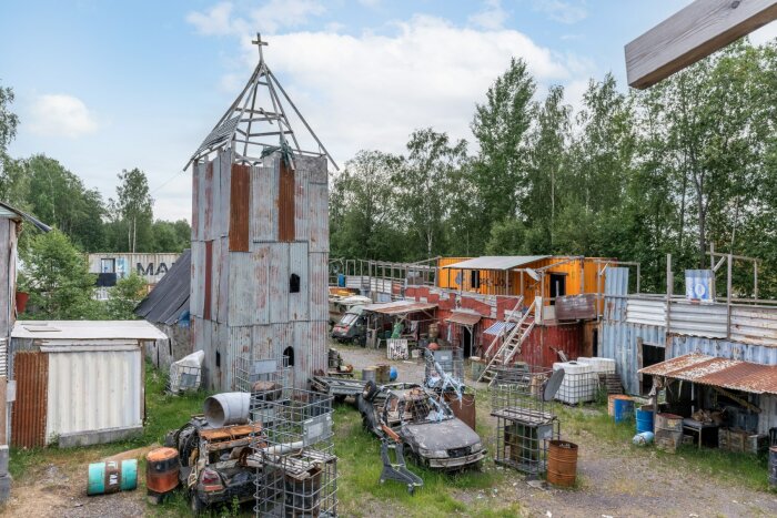 Övergiven plats, improviserat kyrktorn, skrot, containerbyggnader, förfall, grönska, molnig himmel.