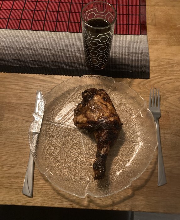 Grillad kycklinglår på en glastallrik med bestick och ett glas mörk dryck på ett rött bordstablett.