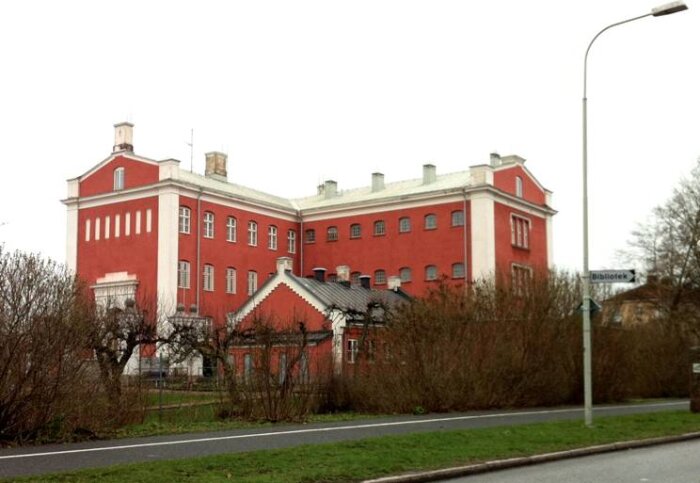 En stor röd byggnad med vita fönster och detaljer, tidigare ett fängelse, med ett busshållplatskylt framför.