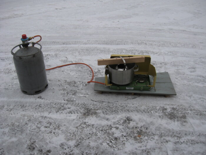 Utomhusbild av smältutrustning för tätmedel bestående av en gasolbrännare och en gryta på ett släde mot en snöig bakgrund.
