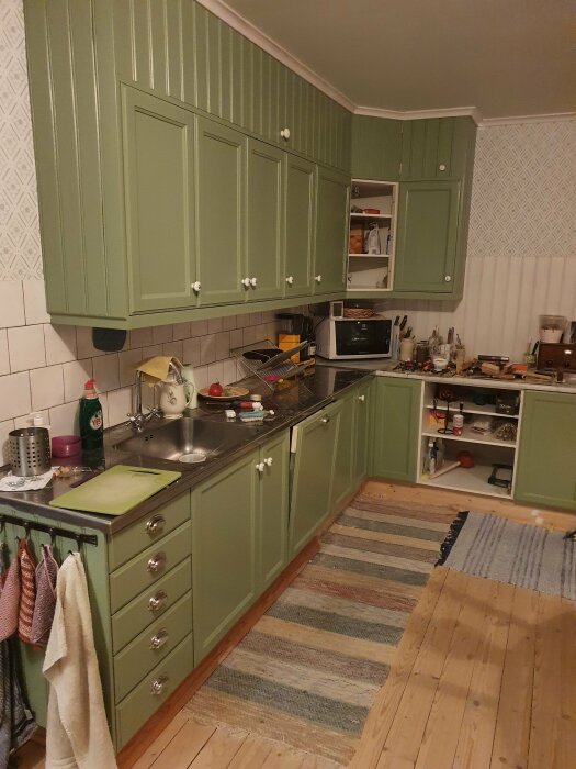 Grönt kök med öppna skåp och diskbänk, använda köksredskap och matta på trägolv.