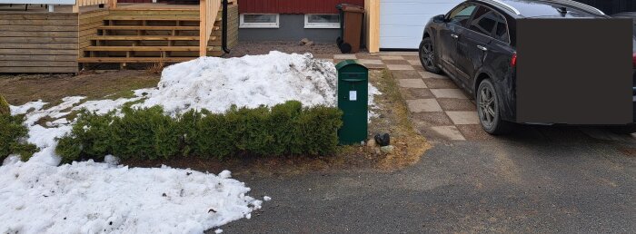 Vår, smältande snö, gröna buskar, brevlåda, bil, garage, trätrappa, stenplattor, bostadshus, grå himmel.