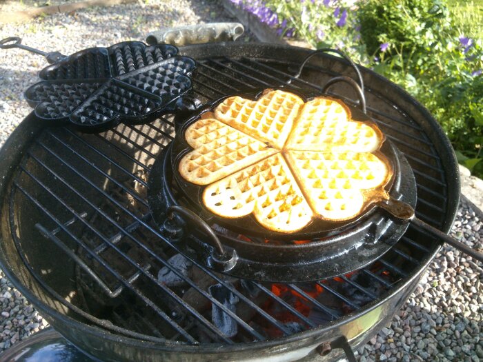 Gjutjärns-våffeljärn på grill med nygräddad våffla, mot bakgrund av blommor.