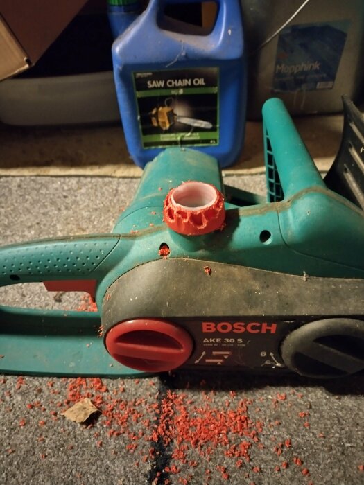 Bosch elektrisk motorsåg på ett golv med röda gnagda plastbitar och saknat lock för kedjeolja.