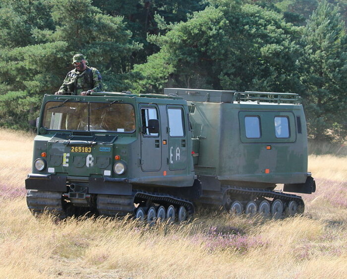 Militär bandvagn med soldat på taket i terrängen, liknar en motoriserad husvagn.