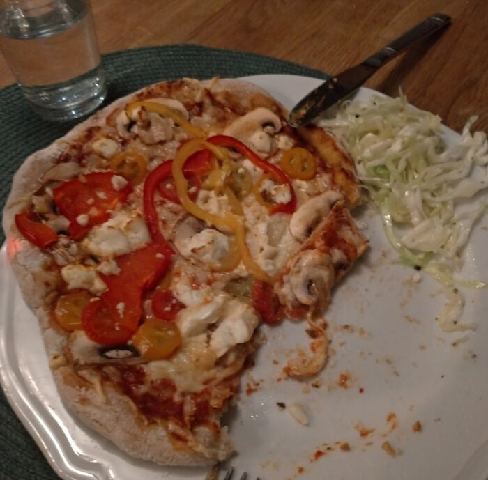 Halväten hemlagad pizza med paprika, champinjoner och ost på en tallrik bredvid en sallad.