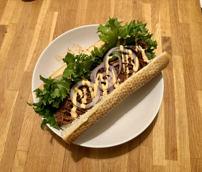 Baguette med pulled beef, grönsallad, rödlök och dressing på en vit tallrik.