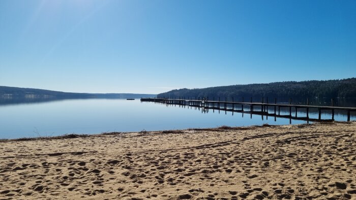 En sandstrand som leder till en brygga och en lugn sjö omgiven av skog och klar himmel.