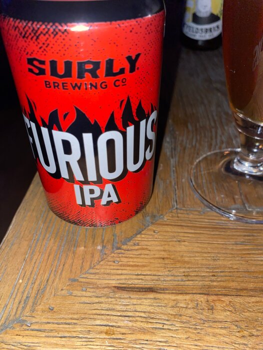 En ölburk märkt "FURIOUS IPA" från Surly Brewing Co, på ett träbord, med glas i bakgrunden.