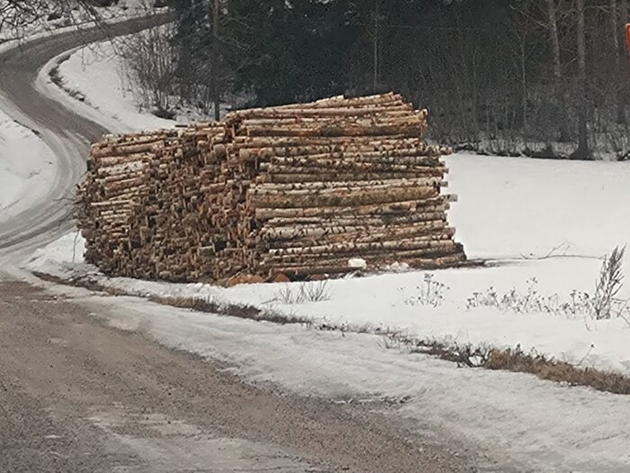 Stapel timmerstockar vid en snöig vägsväng i skogen.