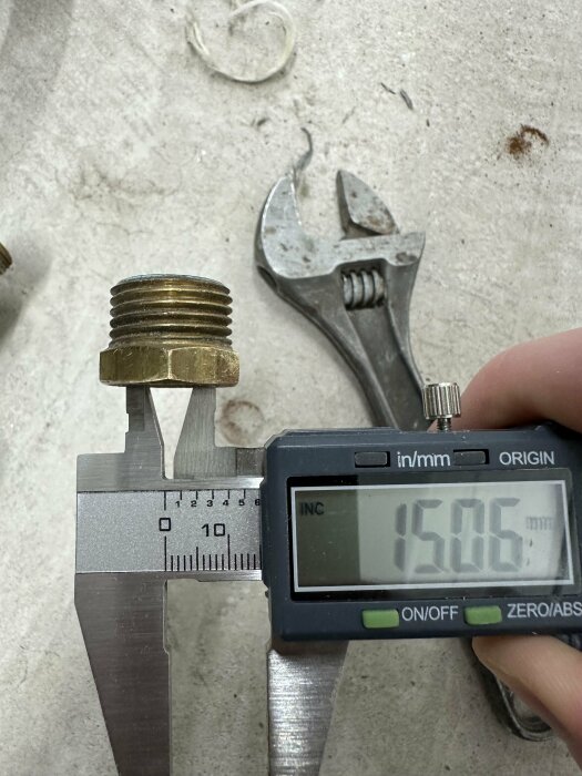 Digital skjutmått mäter metallgängad del framför rostig skiftnyckel på stenytan.