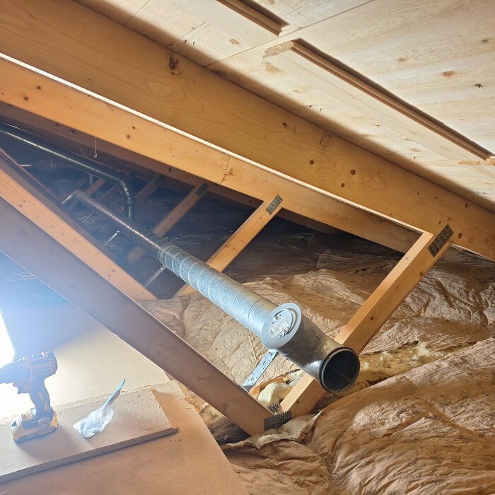 Vind med träbjälkar, isolering, ventilationsrör, byggmaterial och ett batteridrivet verktyg på golvet.