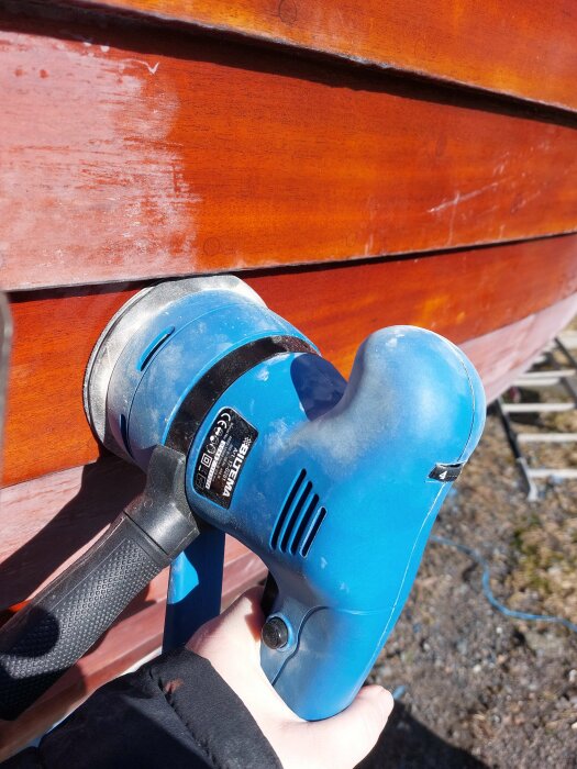 En blå elektrisk slipmaskin används för att slipa en röd träyta utomhus.