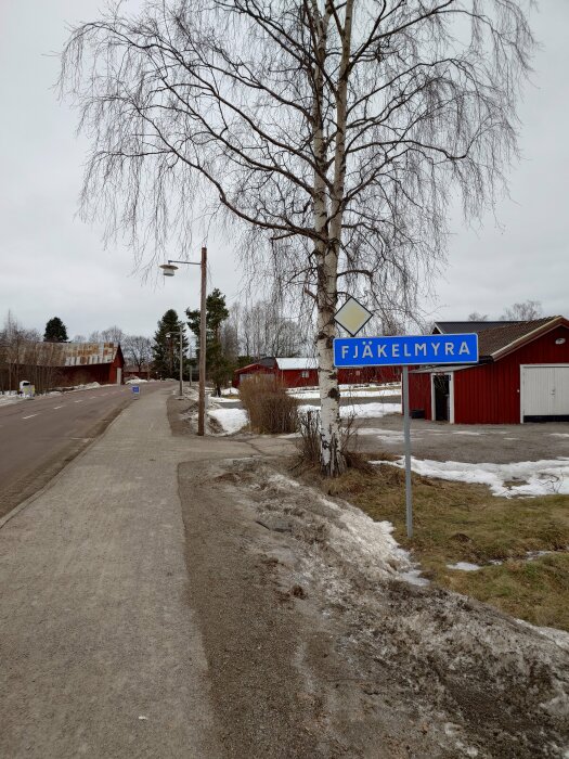 Vägskylt med texten 'FJÄKELMYRA' vid en trottoar med snökanter och en björk i övergången mellan vinter och vår.