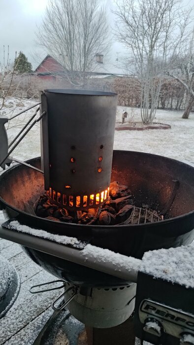 Eldning i en skorstenständare på en grill över snötäckt mark med vinterträdgård i bakgrunden.
