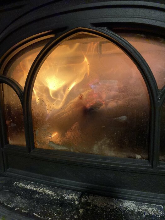 Brinnande ved i en svart gjutjärnskamin med delvis sotigt glas.