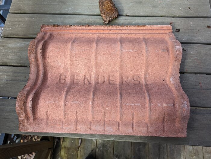 Tegelröd takpanna från Benders på ett träbord, märkt med "BENDERS" i relief.