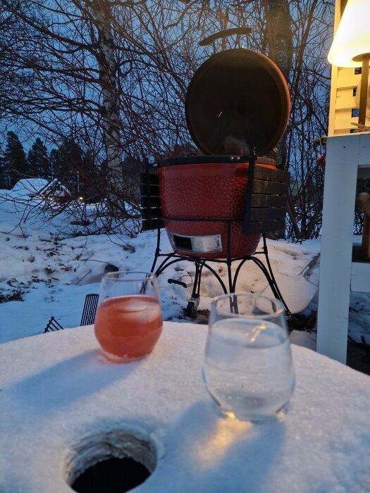 Grill med öppet lock i en snöig trädgård vid skymningen, två glas med dryck i förgrunden.