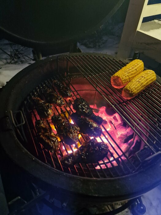 Grillade kycklingben och majs på en grill över glödande kol i snöig omgivning.