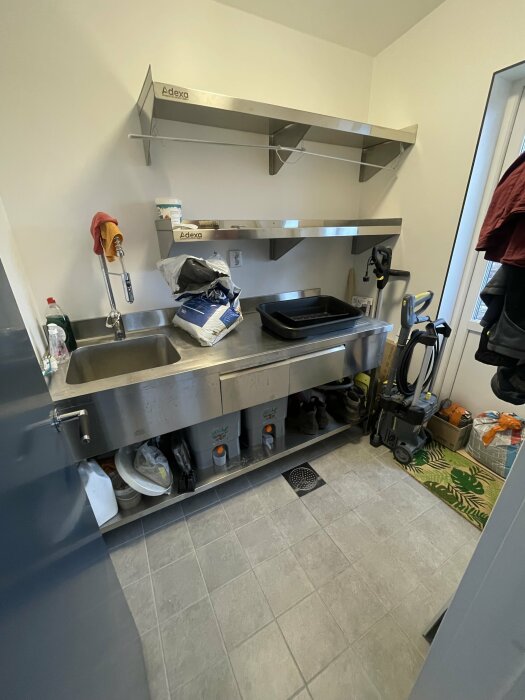 Industriellt kök med diskho, hyllor, rengöringsmedel, städutrustning och oorganiserade föremål på golvet.
