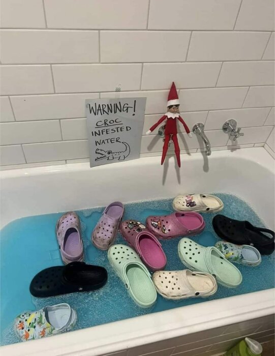 Elf-on-the-Shelf leksak i badrum sitter bredvid skylt som säger "VARIANT! Croc-infesterat vatten" med flera Crocs-skor flytande i badkaret.