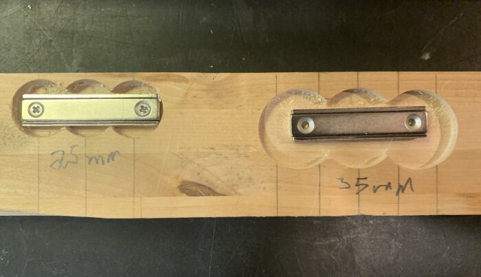 Två magneter på träplanka med utsatta mått 25 mm och 35 mm samt borrningsspår.