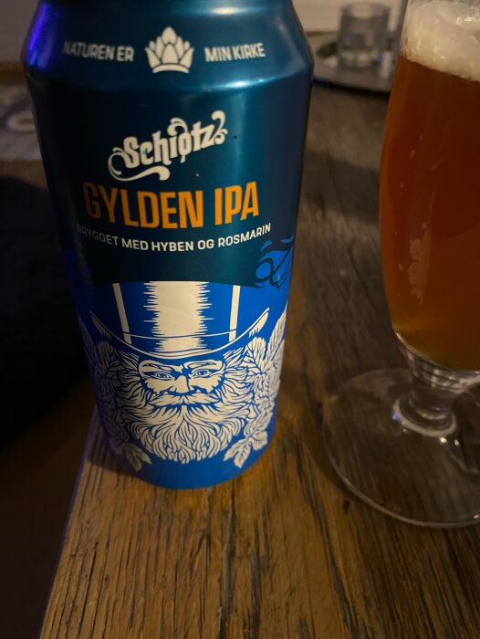 En ölburk med texten "Gylden IPA" och en skäggig figur, bredvid ett glas öl på ett träbord.