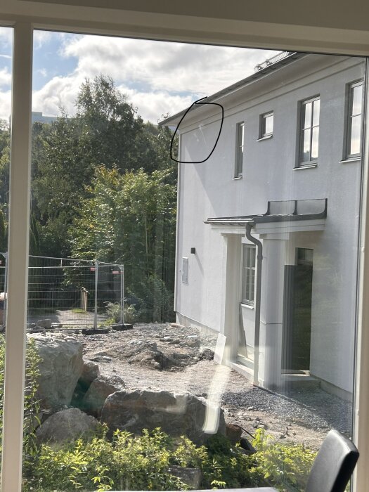Vy genom ett fönster visar en del av ett nybyggt hus med vit putsfasad och stora stenblock i trädgården.
