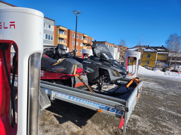 Elbil som laddas vid Tesla Supercharger med ett släp med två snöskotrar parkerat bakom.