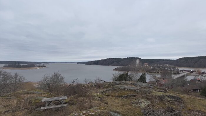 Utsikt över en sjö med en bänk i förgrunden, skog och byggnader i bakgrunden under molnig himmel.
