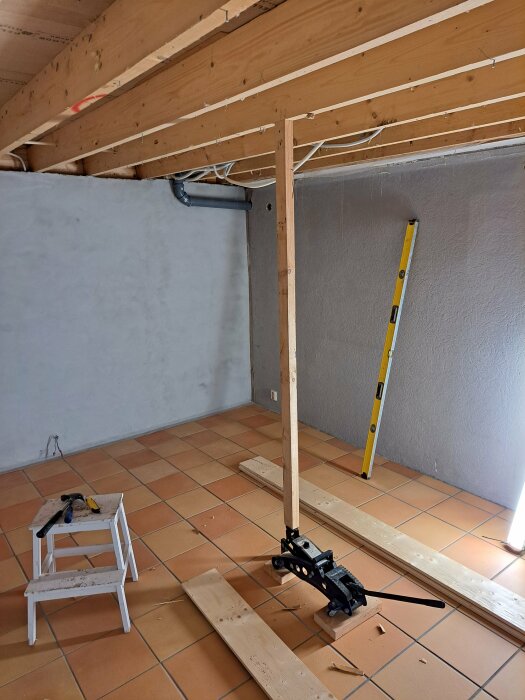 Renoveringsarbete med domkraft, stödben och plywood på ett kaklat golv i ett rum under konstruktion.