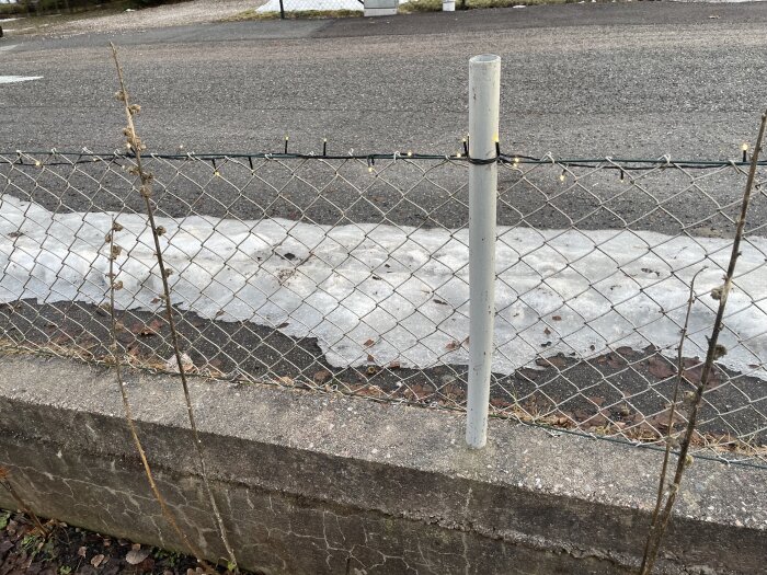 Slitet staket med metallstolpe på en asfalterad yta, viss snö och löv synliga.