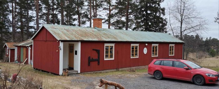 Rött trähus med patinerat tak, skorsten, skogsbakgrund, röd bil framför, landsbygdsmiljö.