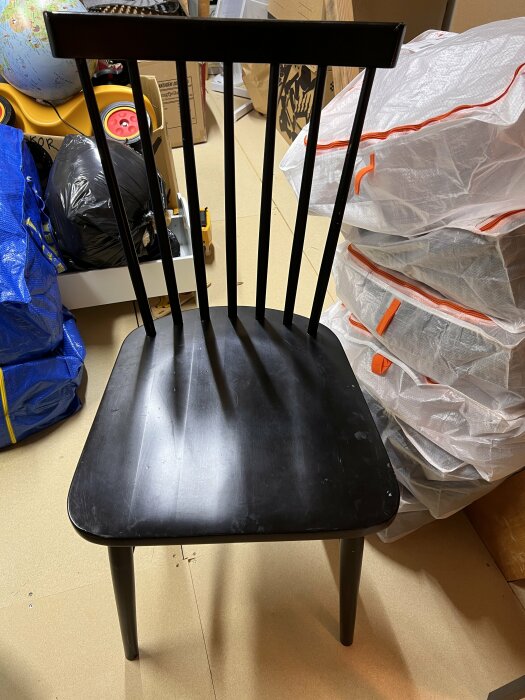 Svart, lackerad stol med raka ryggpinnar och sliten sits, placerad i en rörig vindsmiljö.
