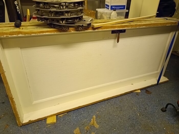 En vit, liggande innerdörr med paneler, som stöds av träbitar och tejp, förberedd för renovering.