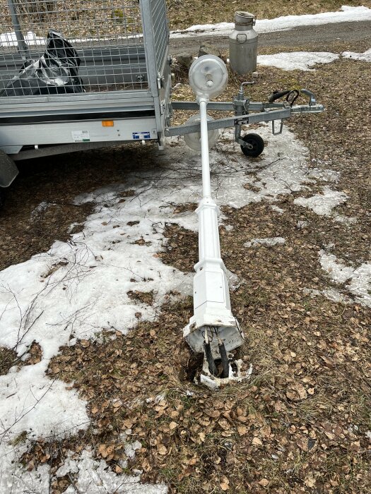 Nedvälvd gårdspelykta med trasigt fäste mot marken bredvid en släpvagn på en grusig yta med snöfläckar.