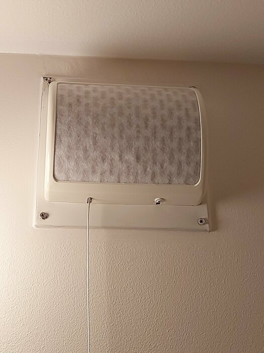 Äldre vit ventilationskåpa med tygfilter och snöre för timer ovanför en köksvägg.