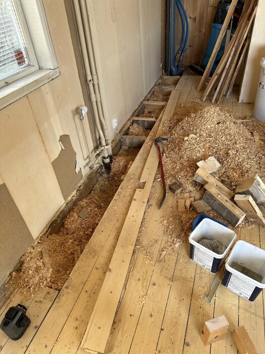 Renovering pågår, upprivet golv, spån, rörledningar, byggmaterial och verktyg syns.