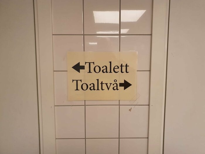 Skylt på kakelvägg med pil vänster "Toalett" och pil höger "Toaltvå" mot vit bakgrund.