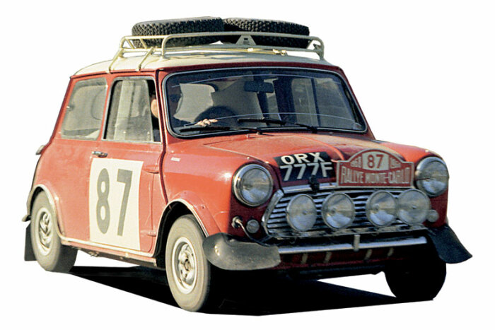 Röd klassisk Mini Cooper med startnummer 87 och extra stora rallyljus fram, samt ett reservdäck monterat på takräcket.