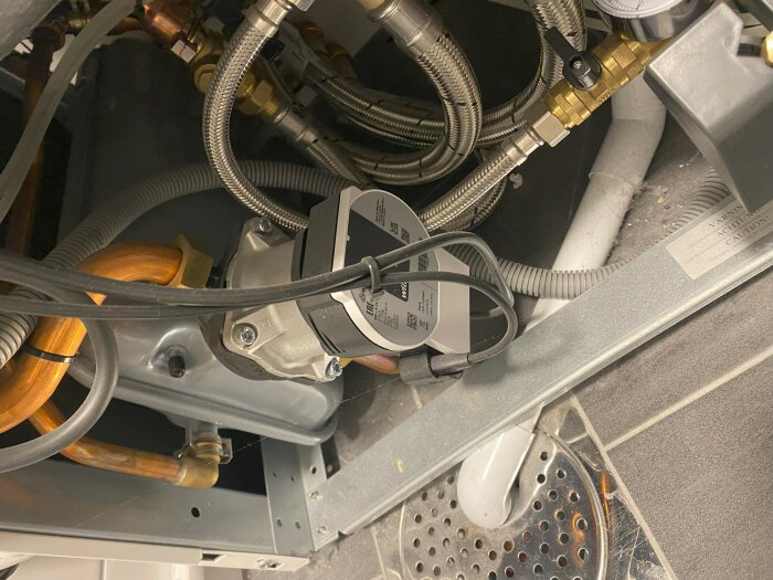 Interiör av en maskin med synliga kopparledningar, slangar och en elektrisk motor, allt placerat ovanför ett ventilationsgaller.