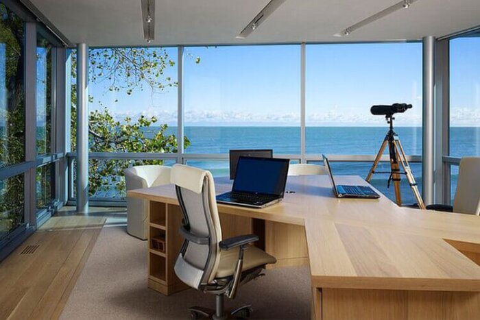 Modernt arbetsrum med stort skrivbord, dator och havsutsikt genom panoramafönster.