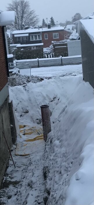 Urgrävning för garageuppfart täckt med snö, synlig brunnsring och gul markduk, omgiven av hus och staket.