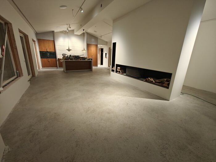 Slipat betonggolv i rum under renovering med kök i bakgrunden och modern öppen spis.