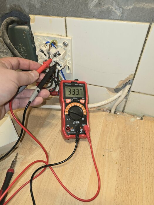 Person mäter spänning i ett eluttag med en multimeter som visar 233,1 volt.