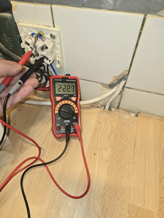 Användning av multimeter för att mäta elektricitet vid öppet eluttag, med kakelvägg och trägolv i bakgrunden.