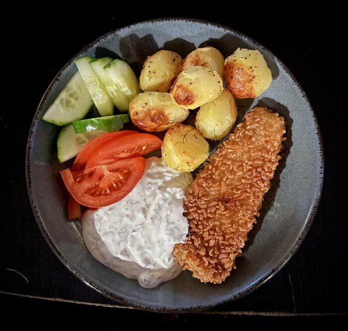 Panerad fisk med ugnstekt potatis, skivad gurka, tomat och hemgjord dillsås på en mörk tallrik.