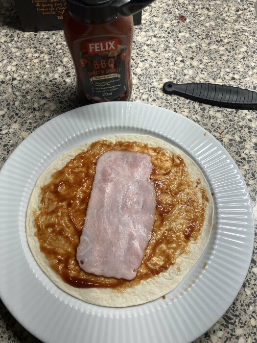 Tortillabröd med bbq-sås, skinka och ost på ett vitt fat redo för brödrosten.