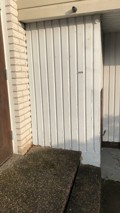 Vitmålad ytterpanel med vertikala brädor bredvid en tegelvägg, nedre del av en utomhustrappa synlig.