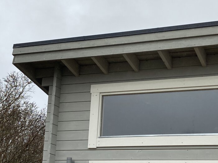 Del av ett nybyggt attefallshus med taköverhang och synliga takåsar, nära ett fönster.